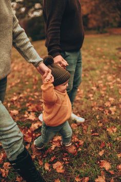 an adult holding the hand of a small child who is walking through leaves on the ground