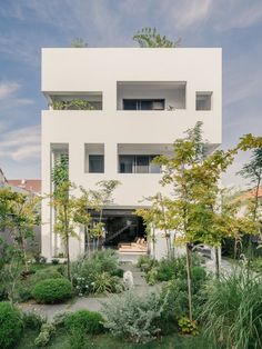 a white building with lots of plants and trees in front of it on a sunny day