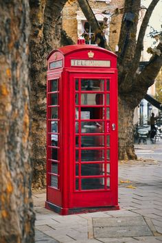 a red phone booth sitting on the side of a road next to a tree and sidewalk