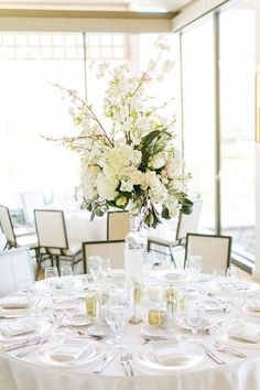 a table set for two with white flowers and place settings in front of large windows