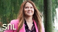 a woman with long hair wearing a pink blazer and black floral shirt standing in front of trees