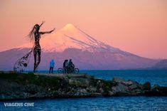 two people standing next to each other near the ocean with bikes in front of a mountain