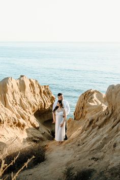 a man and woman standing on top of a sandy beach next to the ocean with rocks