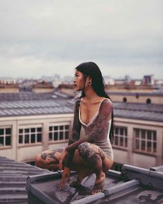 a woman with tattoos sitting on top of a roof looking at the sky and buildings