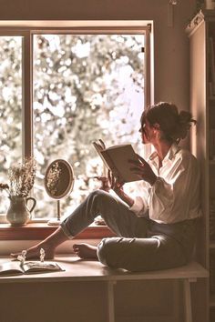 a woman sitting on a window sill reading a book in front of a window