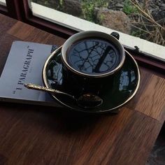 a cup of coffee sitting on top of a wooden table next to a book and window