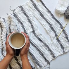 someone holding a cup of coffee on top of a table next to yarn and crochet