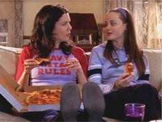 two women sitting on a couch eating pizza and talking to each other in front of them