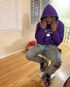 a man sitting on top of a stool wearing a purple hoodie and holding his hands to his face