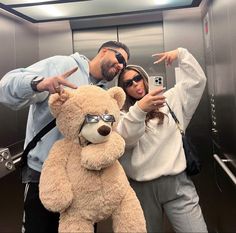 two people taking a selfie in an elevator with a giant teddy bear and sunglasses