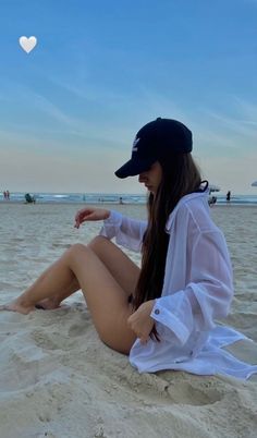 a woman sitting on top of a sandy beach next to the ocean with a heart shaped object in the sky