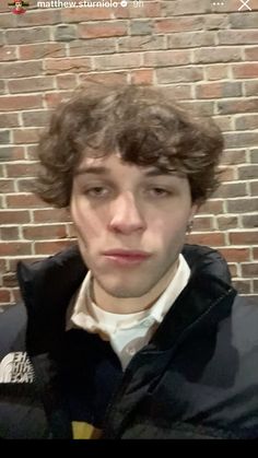 a young man with curly hair wearing a black jacket and white shirt, standing in front of a brick wall