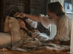 two women in period clothing laying on a bed together, one is brushing the hair of another woman's head