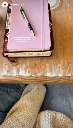 a pink notebook sitting on top of a wooden table next to someone's feet