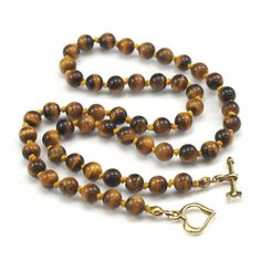 Tiger's Eye Necklace with Gold Plated Toggle Clasp Approx. 18 1/2" long Bead Size : 6mm Strung and knotted on silk cord Handcrafted at Beads of Paradise NYC Tiger’s Eye is a form of Chalcedony quartz, but is what is a known in mineralogy as a pseudomorph. The term comes from the Greek for "false form." Pseudomorphs form when one mineral replaces another. It began as the fibrous blue mineral called Crocidolite, which is made up of iron and sodium. The Crocidolite was gradually transformed into wh Adjustable Lariat Necklace With Gemstone Beads, Gold Jewelry With Hand-knotted Round Beads, Brown Necklace With Round Beads And Lobster Clasp, Adjustable Single Strand Lariat Necklace With Round Beads, Toggle Clasp Necklace With Round Beads, Gold Beaded Necklace With Toggle Clasp, Round Beads Single Strand Lariat Necklace As Gift, Lariat Necklace With Round Beads As Gift, Gold Hand-knotted Necklace For Gift