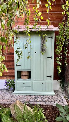 a green cabinet sitting in the middle of a garden next to a brick walkway and trees