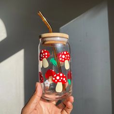 a hand holding a glass jar with red and white mushrooms painted on the inside, in front of a window