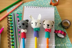 three crocheted animals on top of pencils next to a notepad and markers