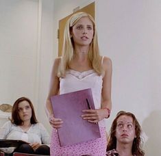 a woman holding a binder standing in front of two women sitting on the floor