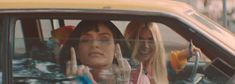 two women sitting in a yellow car making the peace sign with their hands and fingers