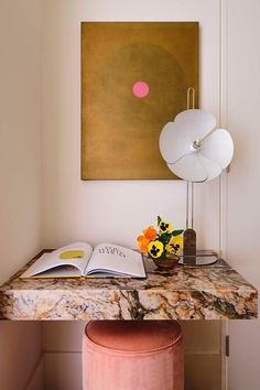 a desk with a book, lamp and flowers on it in front of a painting