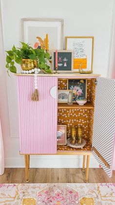 a pink and gold cabinet with pictures on it in the corner next to a potted plant