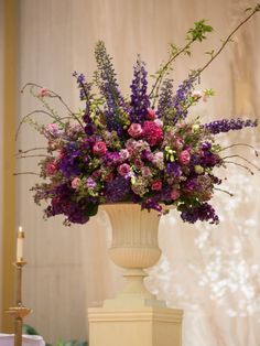 a tall vase filled with lots of purple flowers on top of a table next to a candle