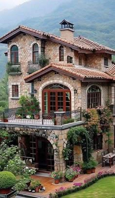 a large stone house with lots of windows and plants on the front porch, surrounded by lush greenery