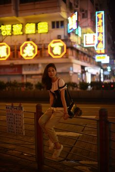 a woman leaning on a fence in front of a building with neon signs behind her