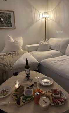 a table with food and drinks on it in front of a white couch, next to a lamp