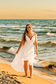 a woman in a white dress is walking on the sand at the beach and smiling