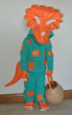 an orange and green dinosaur costume standing next to a ball on a wooden table with a white wall in the background