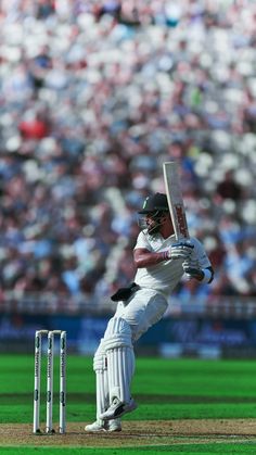 a man in white uniform playing a game of cricket on a field with people watching from the stands