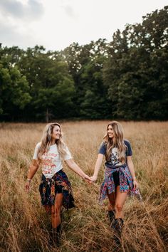 two young women holding hands and walking through tall grass in the middle of a field