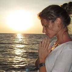 a woman standing in front of the ocean with her hands clasped to her chest as the sun sets