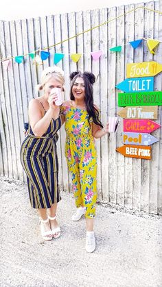 two women standing next to each other in front of a wooden fence with signs on it