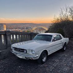 an old white mustang parked on the side of a stone road in front of a cityscape