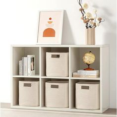 a white bookcase with baskets and pictures on the wall above it, next to a vase filled with flowers