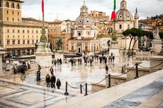 people are walking around in the middle of a plaza with flags on poles and buildings behind them