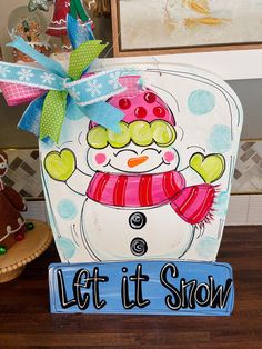 a wooden sign that says let it snow with a snowman in the middle and a bow on top