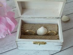 two gold rings sitting in a white box next to a pink flower and seashell