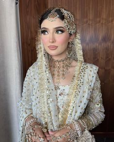 a woman wearing a white and gold bridal outfit with jewelry on her head, standing in front of a wooden wall