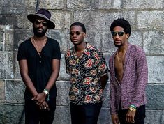 three men standing next to each other in front of a stone wall wearing hats and sunglasses