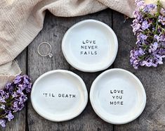 three white dishes with words written on them next to purple flowers and a ring holder