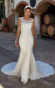 a woman in a white wedding dress standing on a stone patio with barrels behind her