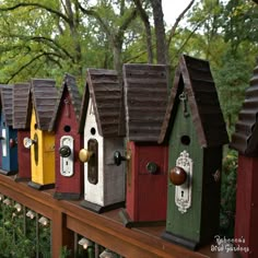 a row of colorful bird houses sitting on top of a wooden fence next to trees