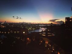 the sun is setting over a city with lights on it's buildings and water in the background