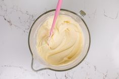 a mixing bowl filled with cream on top of a white marble countertop next to a pink plastic spoon