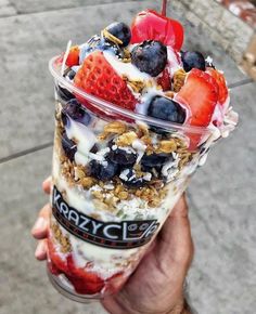 a person holding up a plastic cup filled with yogurt and fruit on top