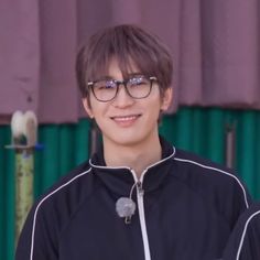 a young man wearing glasses standing in front of a green and purple curtained background